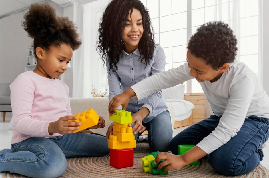 Une ATSEM accompagne 2 enfants à réussir la construction d'un jeu de legos. 