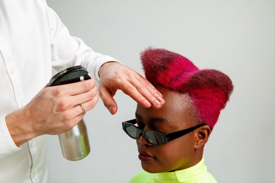 Une jeune femme africaine aux cheveux teint en rouge, habillée d'une jaune éclatant, se laisse coiffer par un coiffeur professionnel tenant à la main 
        à la main droite un aérosol et avec la main gauche posée sur le front de cette jeune femme.